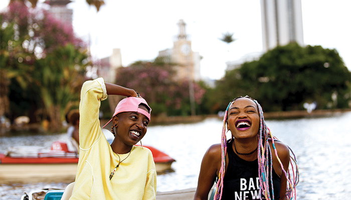 Two women sit on a boat near the edge of a small lake as they smile and laugh