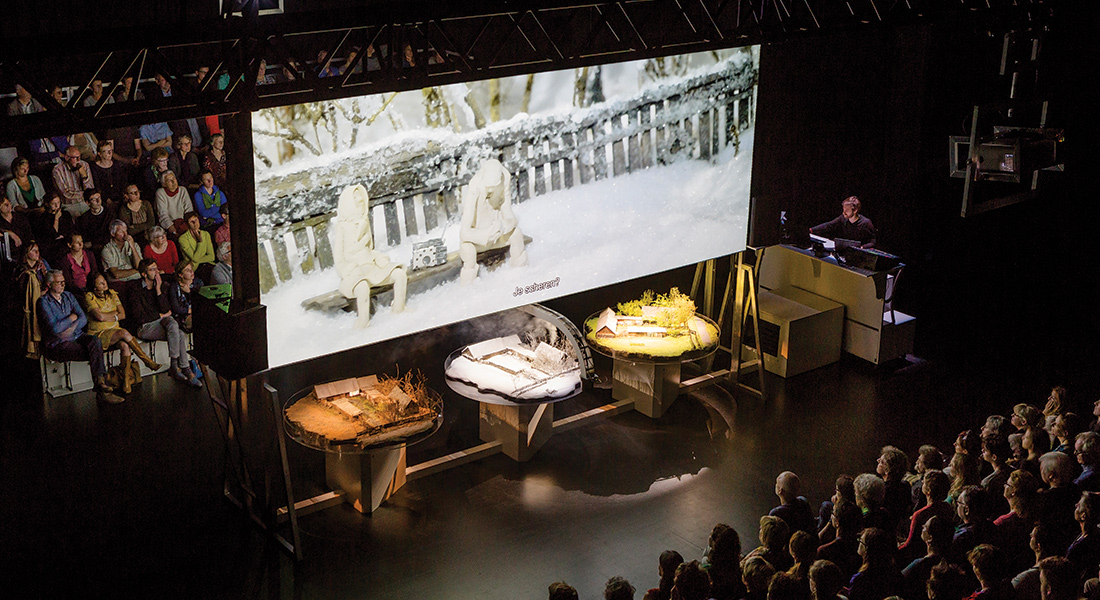 An audience is split into two groups, a large movie screen between them holds an image from a documentary film, and tables beneath the screen hold dioramas in a production still from Zvizdal [Chernobyl - so far so close] by Belgian performance group Berlin
