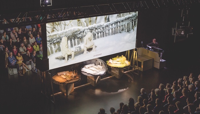 An audience is split into two groups, a large movie screen between them holds an image from a documentary film, and tables beneath the screen hold dioramas in a production still from Zvizdal [Chernobyl - so far so close] by Belgian performance group Berlin