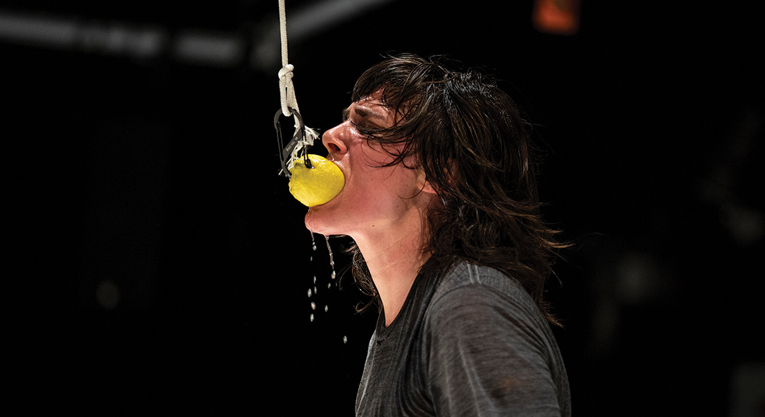 Dance artist Faye Driscoll, seen from shoulders up in a black t-shirt, bites into a lemon hanging from a rope and hook against a dark background