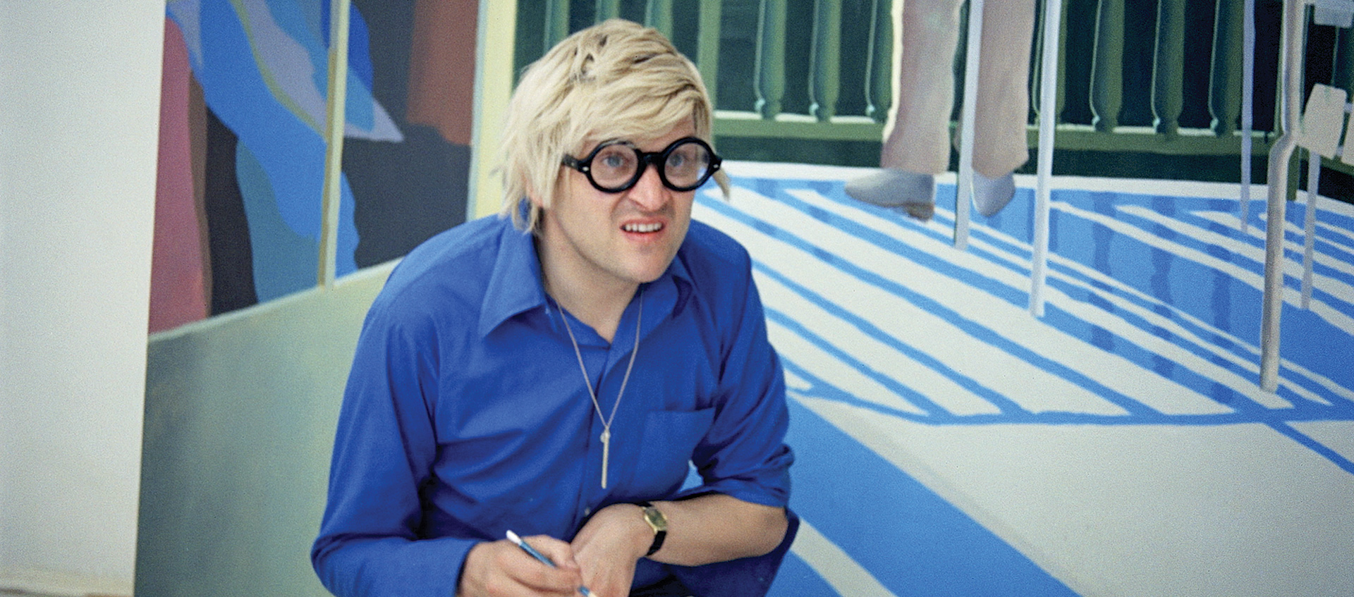 An image of British artist David Hockney sitting in front of one of his paintings in a scene from Jack Hazan's 1973 film A Bigger Splash