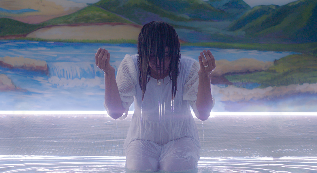 Actor Dira Paes kneels in a shallow body of water, hands up and head bowed with her hair covering her face, as if in deep prayer, in a scene from Gabriel Mascaro's Divine Love