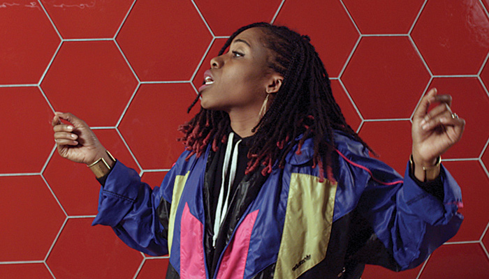 A young woman of color in bright robes sings against a red tile backdrop in the video work RISE by Barbara Wagner and Benjamin de Burca 