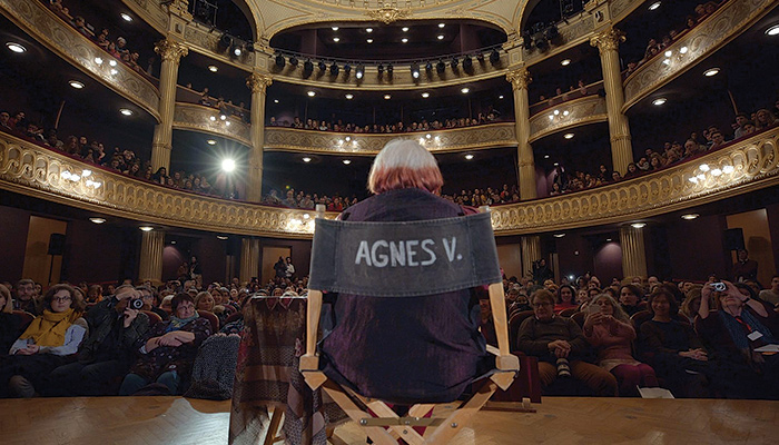 Filmmaker Agnés Varda is seen from behind, sitting in a director's chair with her name on it, on a stage in front of a full audience in a scene from the film Varda by Agnés