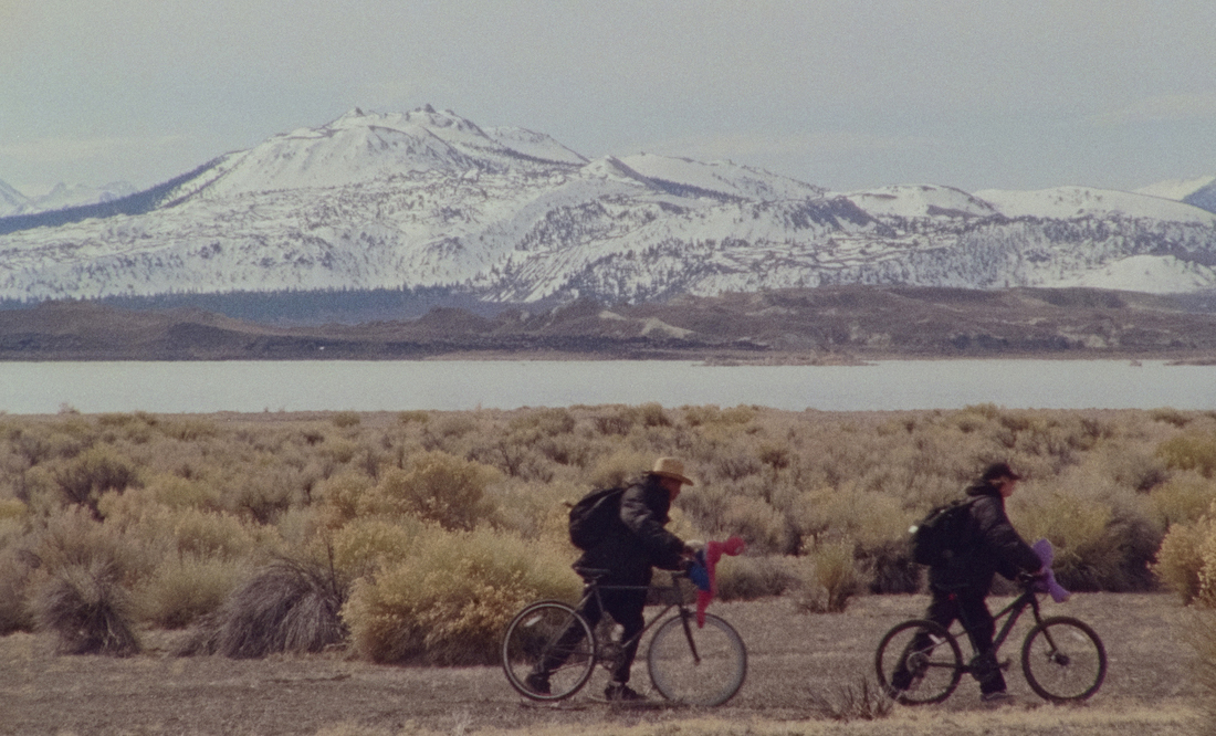 Two teenage boys ride bikes in a grassy, isolated landscape with mountains in the background in a scene from artist Stanya Kahn's short film No Go Backs