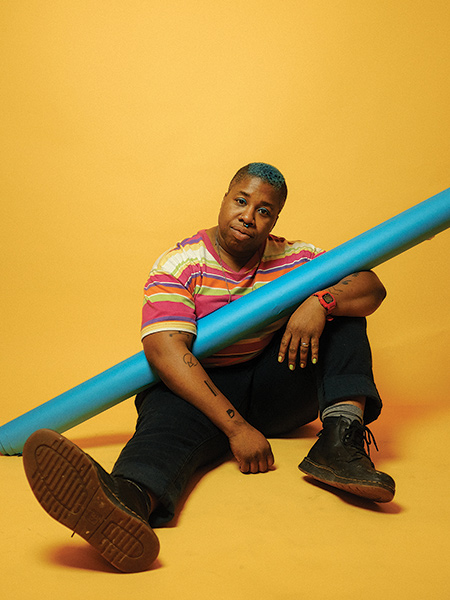 Singer Sharon Udoh sits against a solid yellow background holding a pool noodle that's almost the same color blue as her short afro. Photo by Kate Sweeney