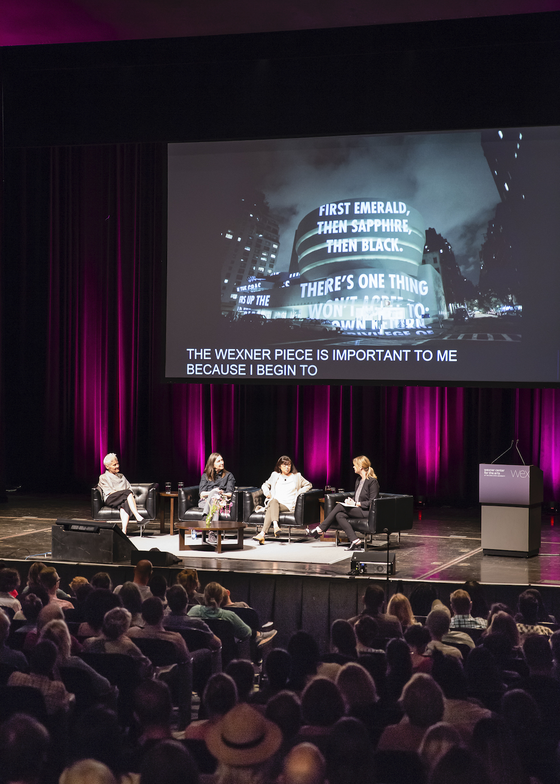 Artists Ann Hamilton, Jenny Holzer, and Maya Lin sitting on the stage of Mershon Auditorium beneath a screen with an image of Holzer's work and closed captioning. The women are discussing their work as part of opening events for the fall 2019 exhibition HERE at the Wexner Center for the Arts. Photo by Katie Gentry