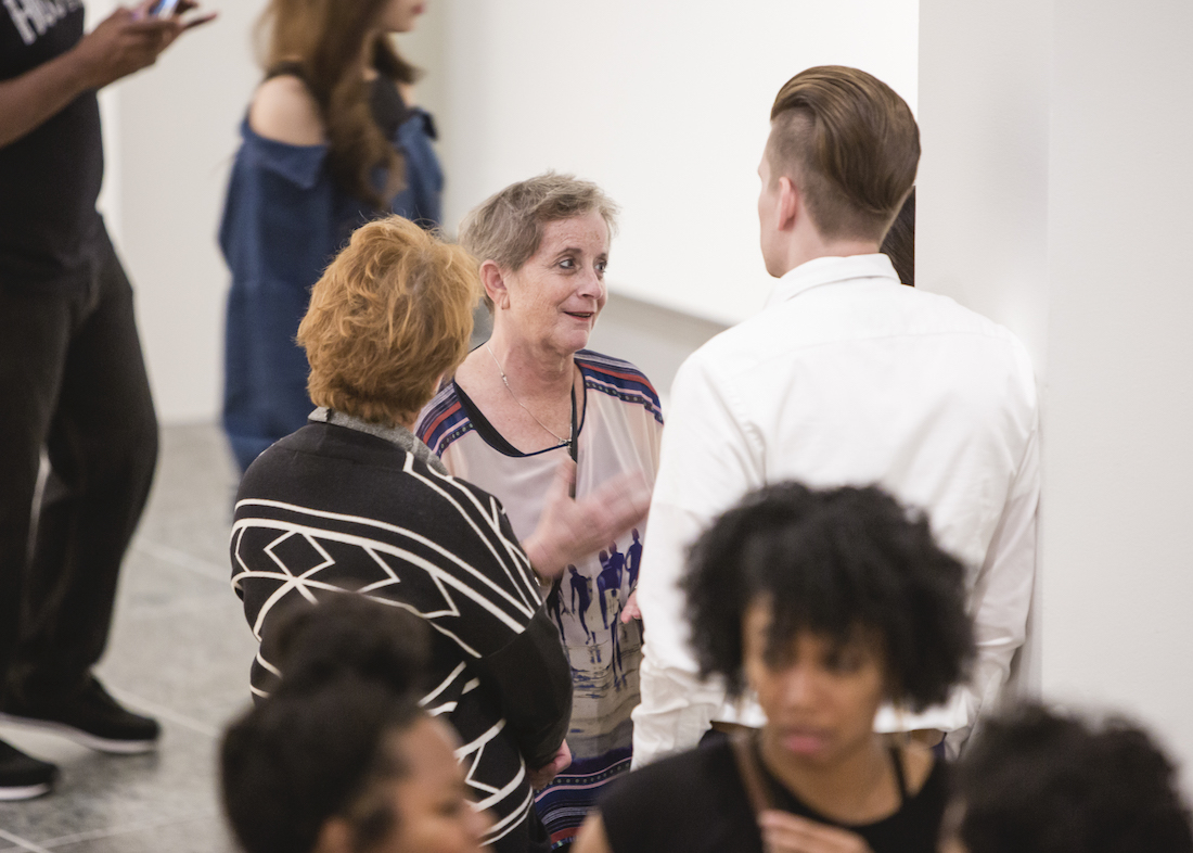 Wexner Center supporter Janice Roth speaks with two others with their backs to the camera in a tight shot of a gallery space at the Wexner Center for the Arts during the opening of the 2018 exhibition Mickalene Thomas: I Can't See You Without Me