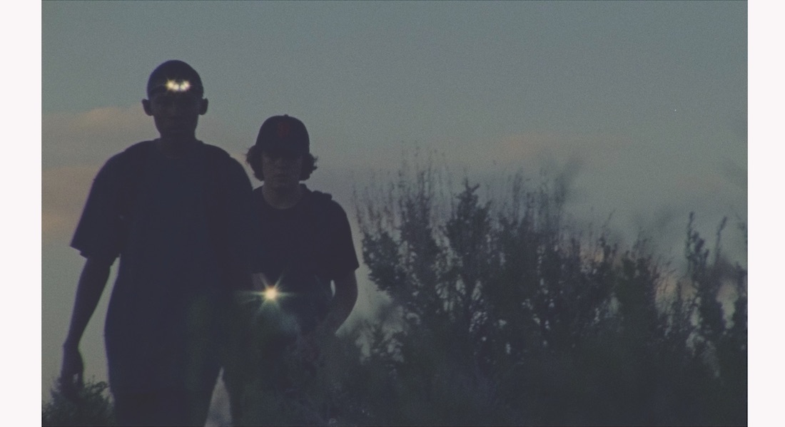 Two young men in baseball caps are seen from behind outdoors at dusk in a scene from Stanya Kahn's short film No Go Backs