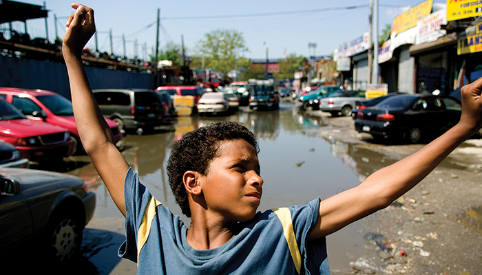 A young boy of color seen from the chest up standing in an auto salvage yard, holding his arms up and outstretched, in a scene from Ramin Bahrani's film Chop Shop