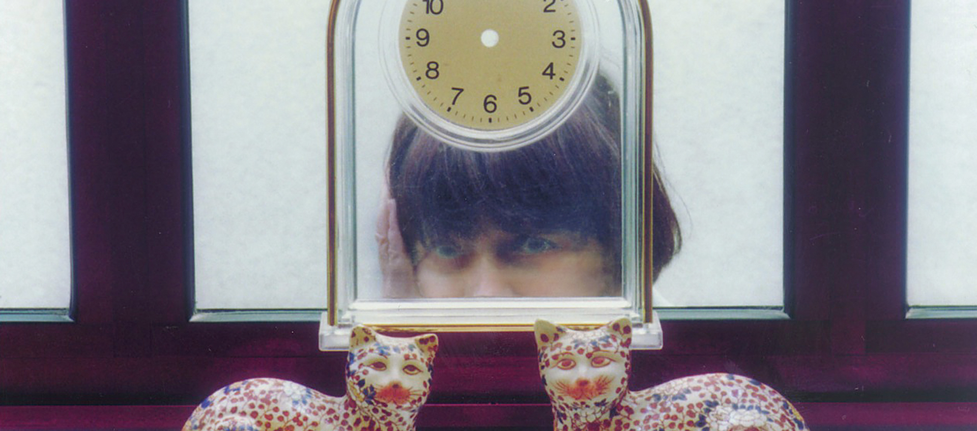 Filmmaker Agnés Varda seen from the cheeks up, standing behind a glass table clock and two decorative ceramic cats