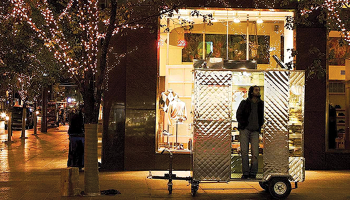 A young Pakistani man stands in front of a food cart outside in the city at nighttime in a scene from Ramin Bahrani's 2005 film Man Push Cart