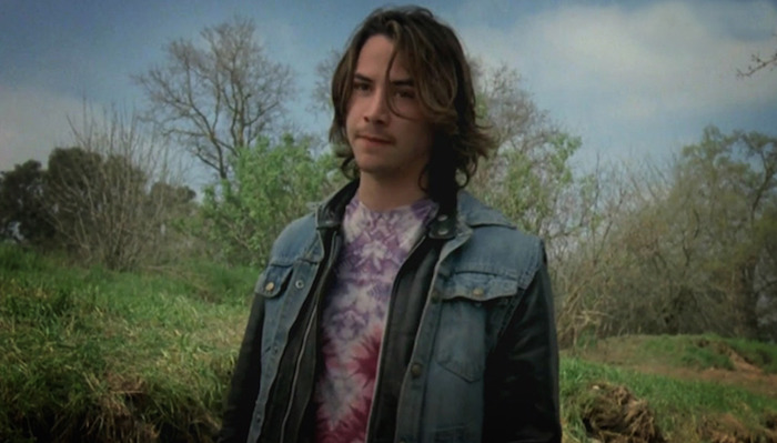 Color image of a young Keanu Reeves standing outdoors in front of grass and trees during the production of Tim Hunter's 1984 film River's Edge