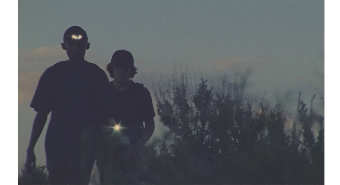 Two teenage boys using flashlights in a brush-filled outdoor area at sunset walk toward the camera in a scene from Stanya Kahn's 2020 short film No Go Backs