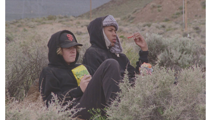 Two teenage boys in hats and coats sit in the California wilderness eating snack food in a scene from Stanya Kahn's 2020 short film No Go Backs