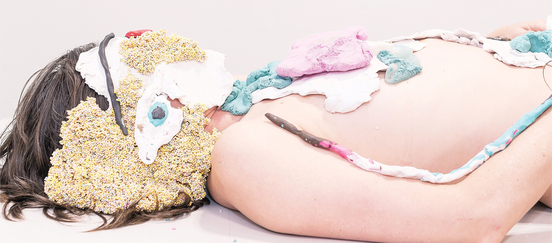 Dancer and choreographer Faye Driscoll from the waist up, lying down on a white surface, her face and parts of her torso covered in thin layers of clay and other materials