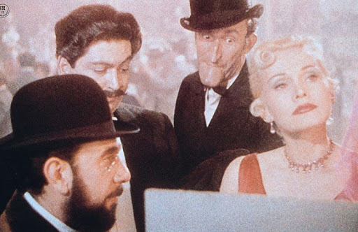 A head-and-shoulders group image of four individuals—three men and one woman—huddling around a poster seen from behind in a scene from John Huston's 1952 film Moulin Rouge