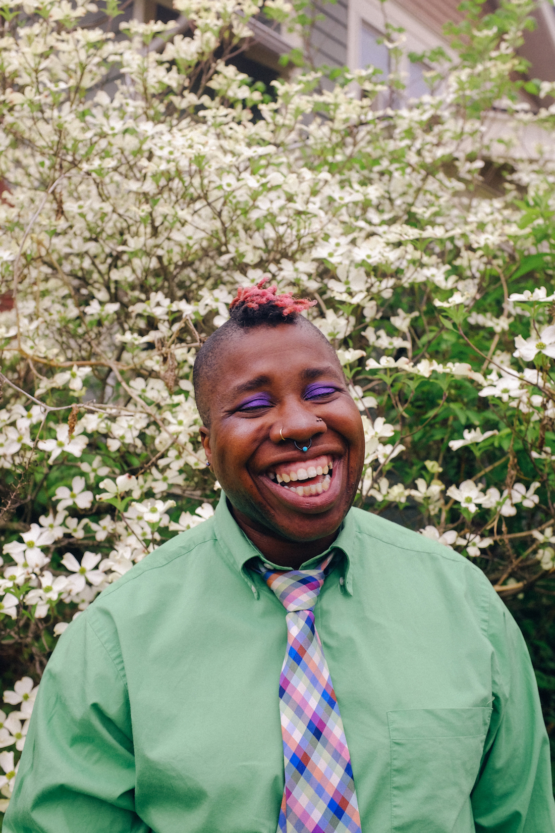 Composer and performer Sharon Udoh, an African American woman with a short colored mohawk wearing a green button-up shirt and a plaid tie, stands smiling in front of a tall bush with white flowers