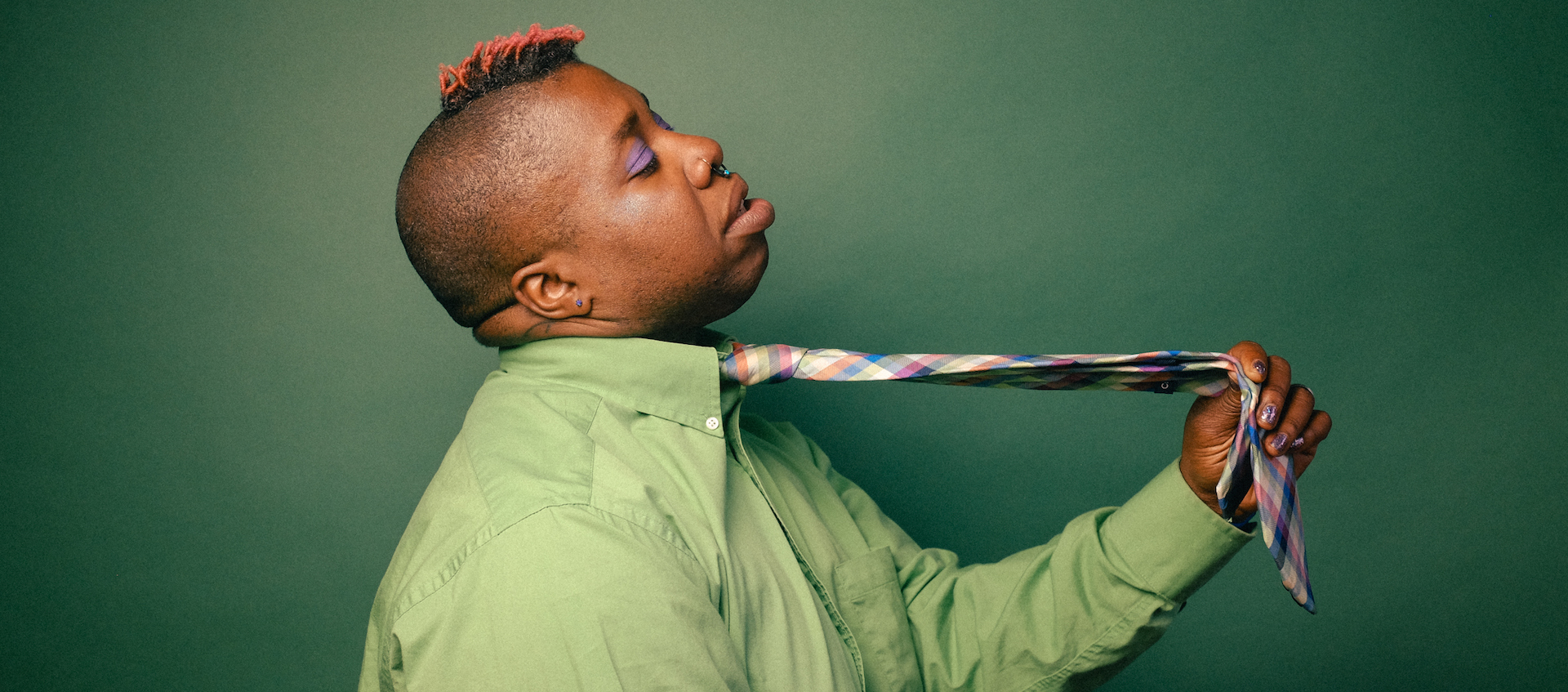Composer and performer Sharon Udoh, an African American woman with a short colored mohawk, stands in profile against a green background, wearing a green button-down shirt and pulling on a tie around her neck