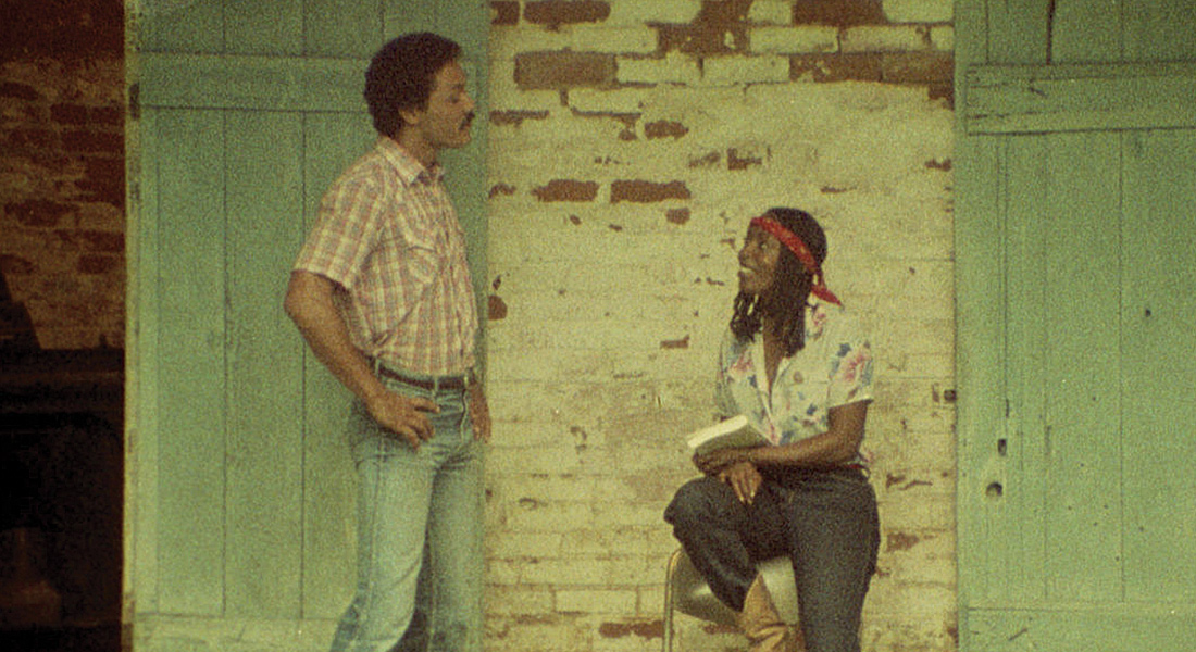 Peter (actor Richard Romain) stands conversing with Maria, (Tommye Myrick) seated on a stool in front of a brick wall. 