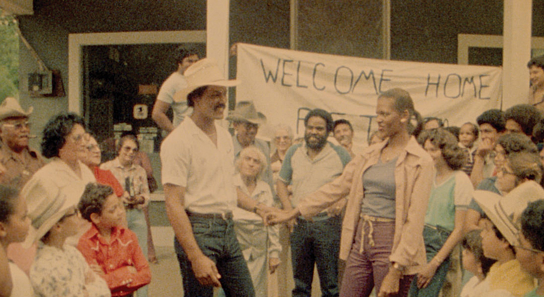A group of people gather around to greet the male protagonist, Peter, with a “Welcome Home” banner in the background.