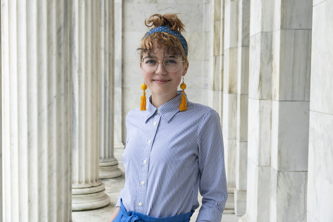 A waist-up color picture of Wexner Center for the Arts intern Kat Arndt, a young white woman with bangs, a top bun, glasses, orange dangly earrings, and a blue button-up shirt.