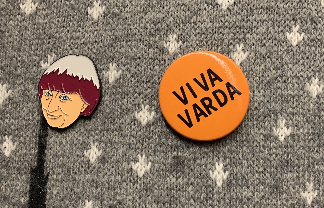A close-up of enamel pins of Agnés Varda's head and the words "Viva Varda" on an orange background, pinned to a gray and white sweater. Pins are from the collection of Wexner Center Associate Film/Video Curator Chris Stults