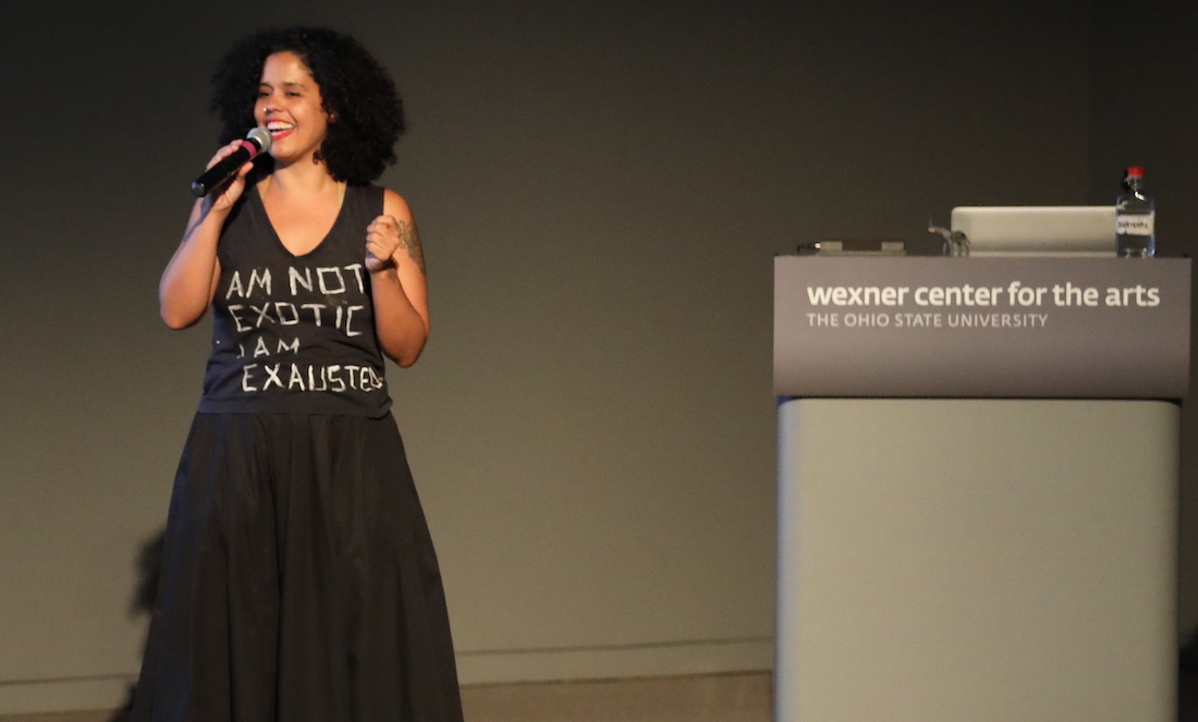 Latinx artist Awilda Rodriguez Lora in a T-shirt that reads "I am not exotic I am exhausted" speaks from the stage of the Film/Video Theater at the Wexner Center for the Arts