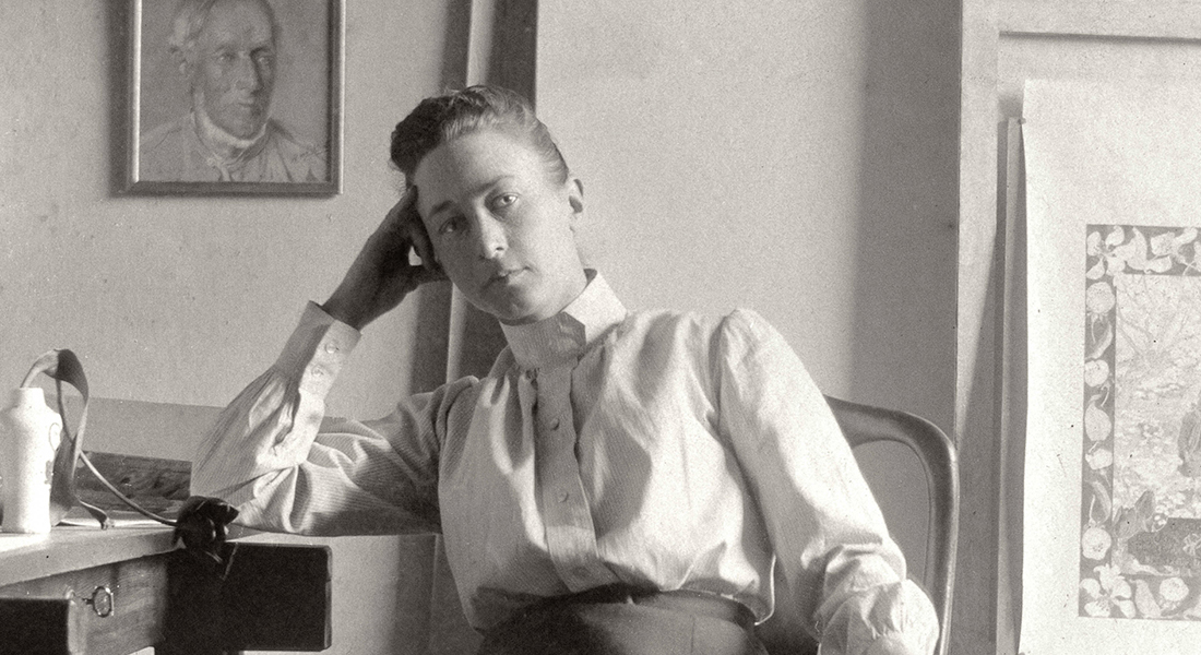 Painter Hilma af Klint in an archival photo posed, sitting on a chair with hand holding head and elbow on desk looking straight at camera