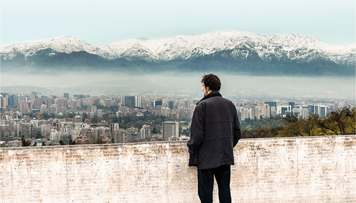 a man in a black coat overlooking a city with mountain in the background