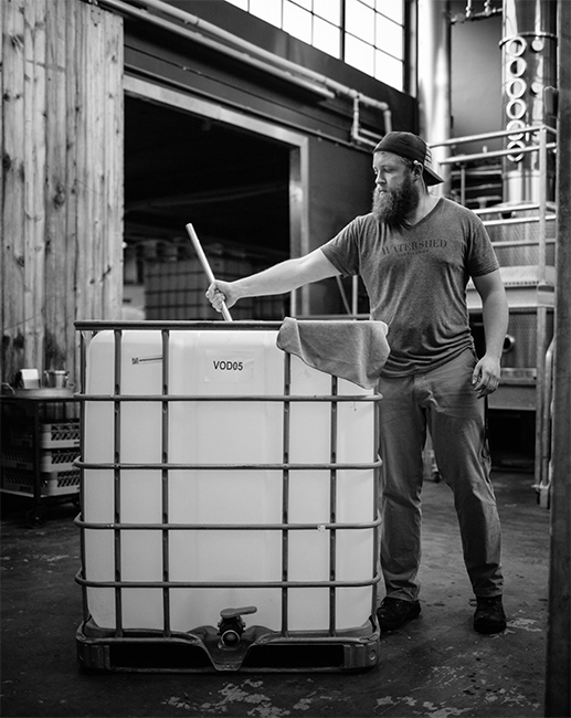 A black and white image of a worker at Columbus' Watershed Distillery mixing a clear plastic vat of hand sanitizer