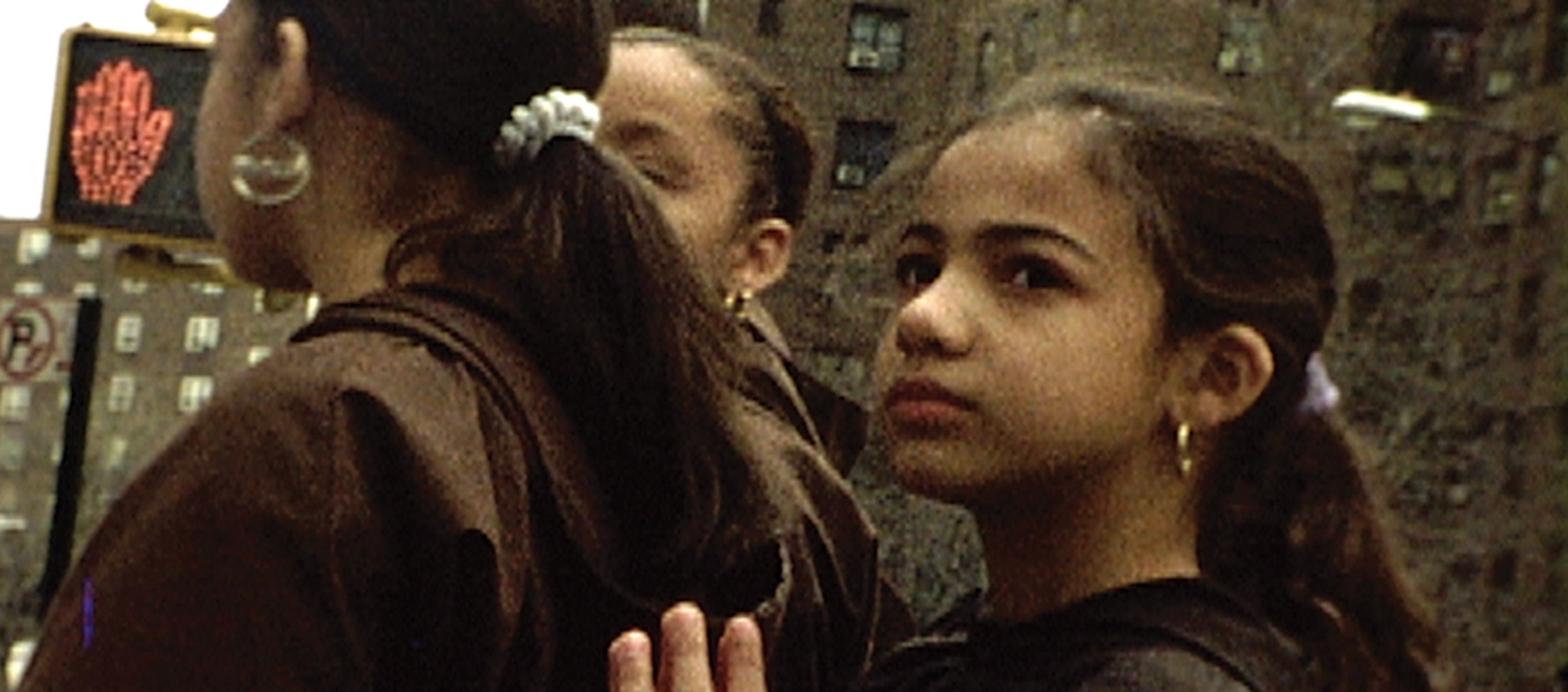 A young girl stands on a street corner behind a woman and looks to the left in a scene from Jeanne Liotta's video work Crosswalk