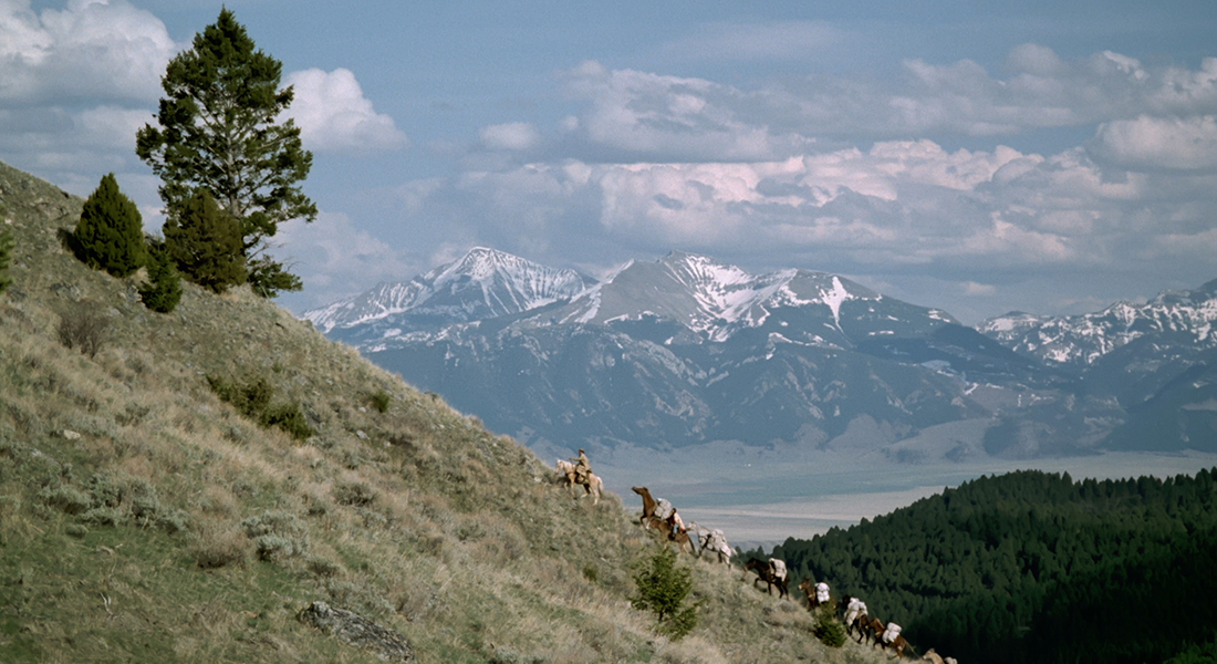 several horses with people and luggage going up a hill with snowy mountains in the background