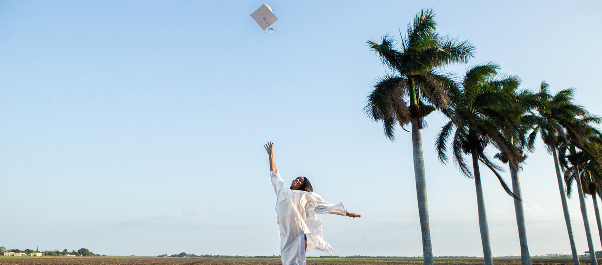 A young woman in a graduation gown throws her cap into the air on a palm tree-lined street in a scene from the documentary Pahokee