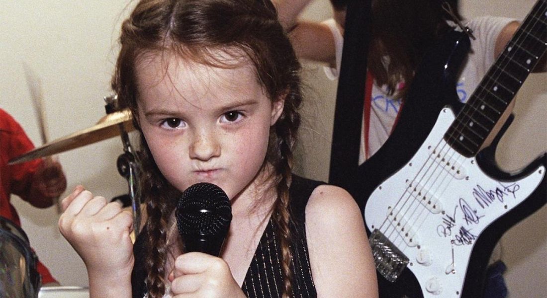 A little white girl with brown braids holds a microphone to her mouth and raises a fist in the air in a scene from the documentary Girls Rock!