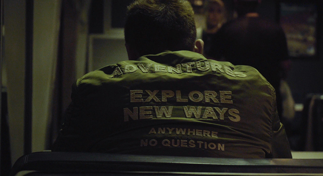 A seated man wearing a jacket with the words 'adventures explore new ways anywhere no questions'