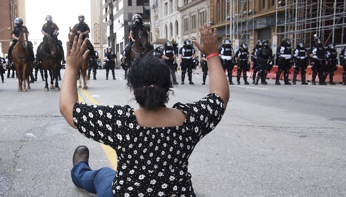 A young woman of color with her back to the camera sits with her arms raised in front of a line of police officers during a protest for the death of George Floyd in Columbus, Ohio, Saturday, May 30, 2020; photo: Mary Barczak
