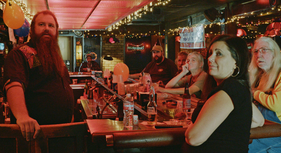 A group of six men and women of various ages sit a dive bar counter tended by a bearded man