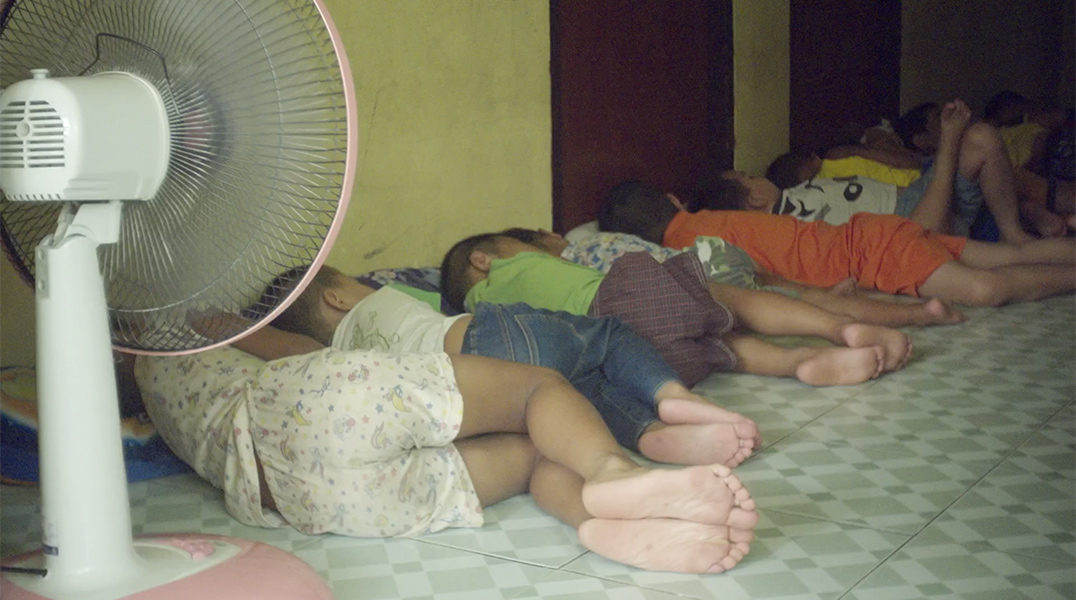 A row of children lay napping on a tile floor with an electric fan pointed at them.
