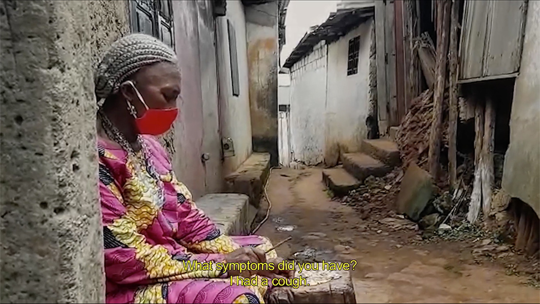 A black woman sits in a doorway wearing a brightly colored dress and a red mask. The subtitled text says "What symptoms did you have? I had a cough."