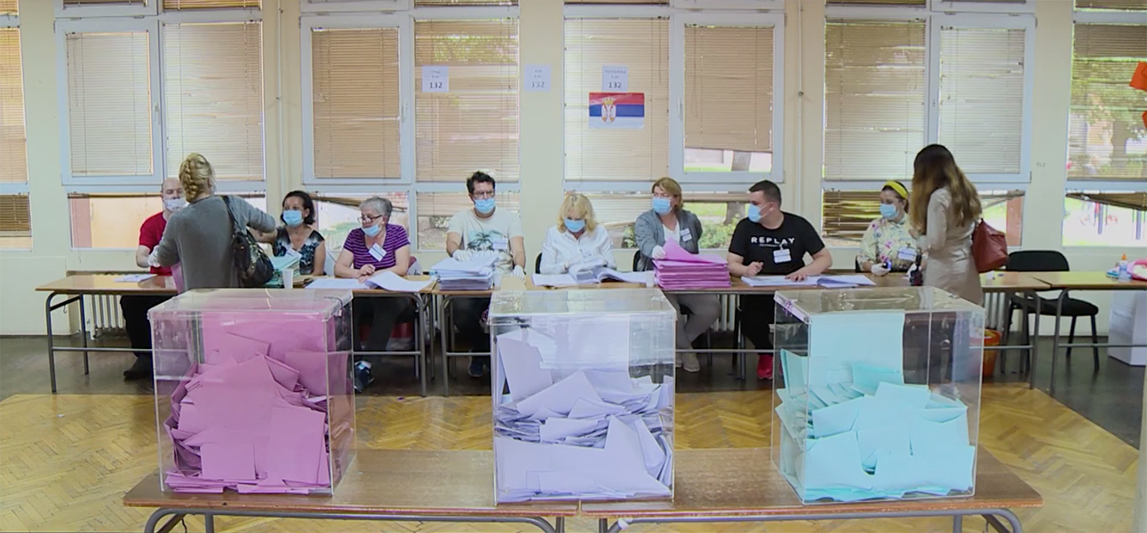 Ballot boxes sit in the foreground with a table full of people wearing disposable masks sits behind them.