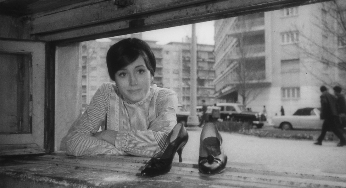 A black-and-white still with Ilda looking through a window next to a pair of high-heeled shoes