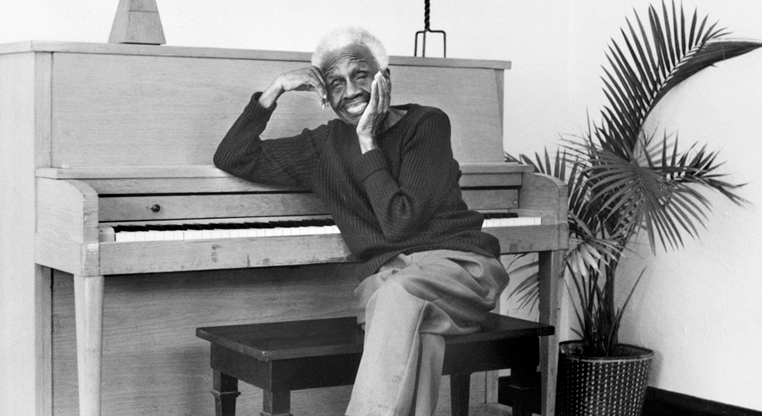 Ruth Ellis sits on a piano bench in front of a piano and houseplant.