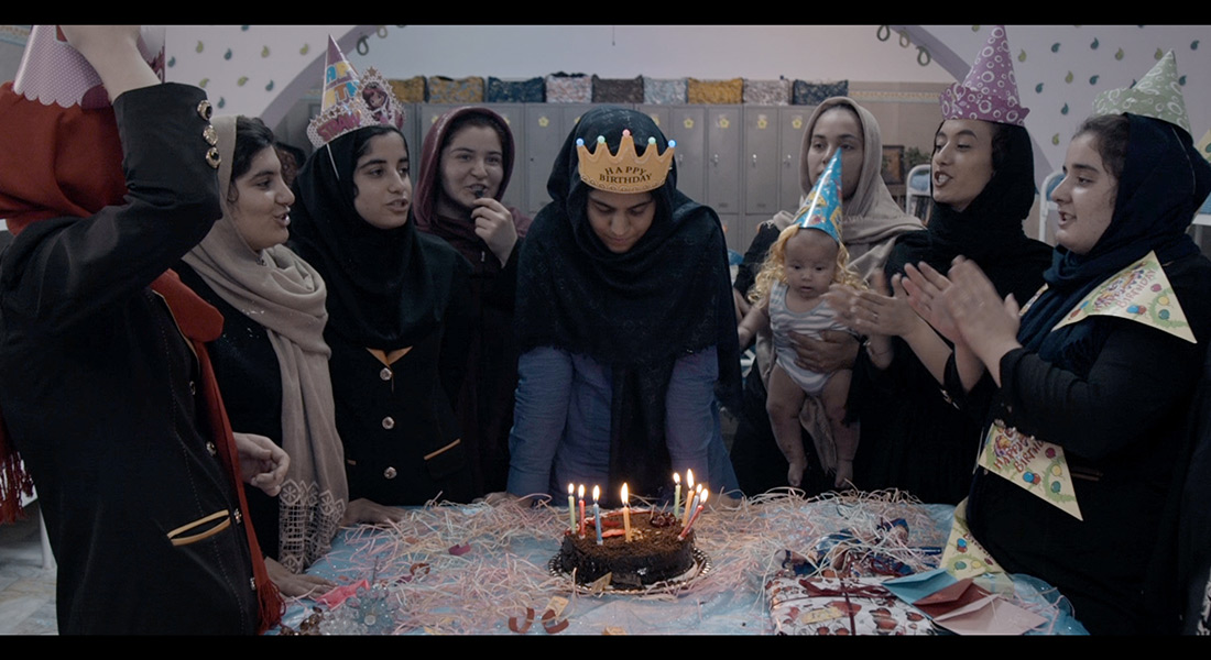 Six young women and an infant stand around a table next to another woman blowing out candles on a birthday cake 