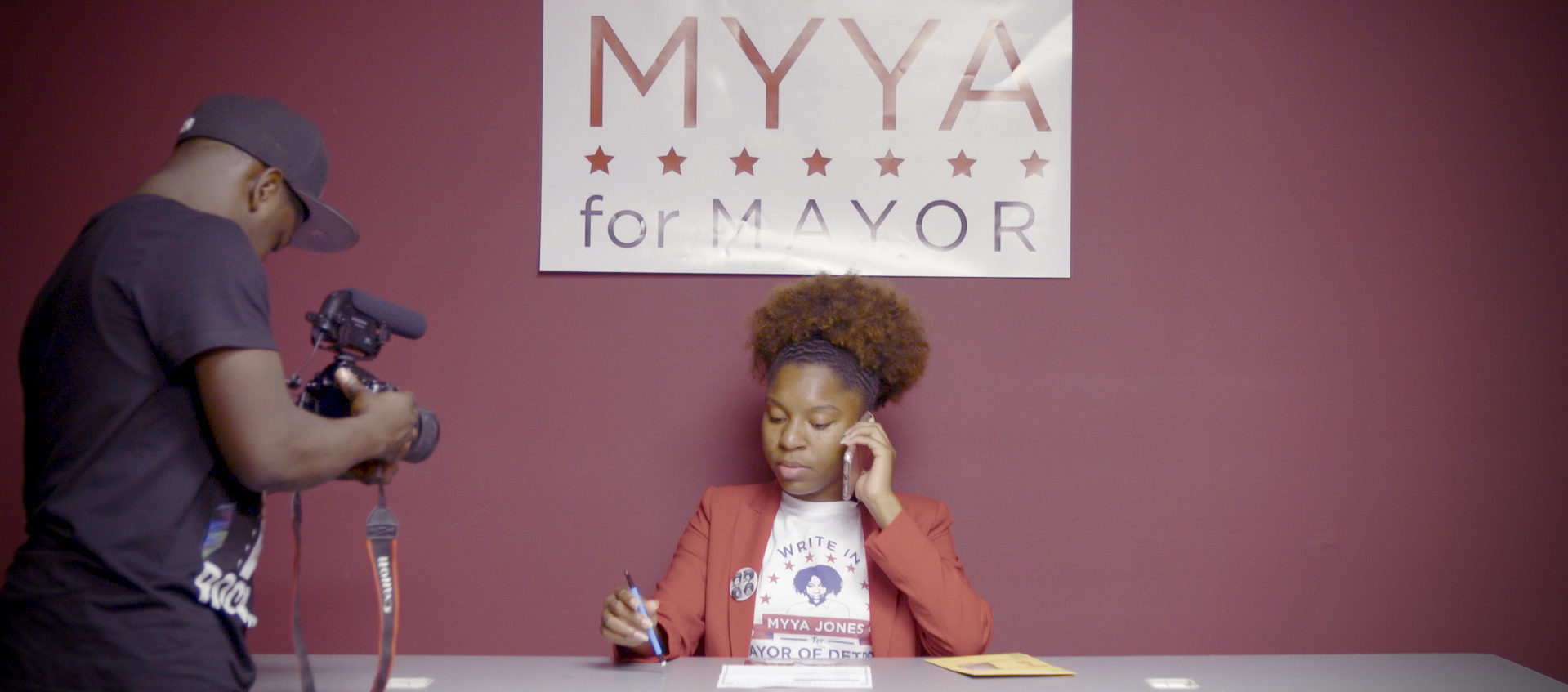 A young black woman mayoral candidate sits at a table on the phone, under a sign for her campaign, as a black man stands to her right, taking her picture with a camera