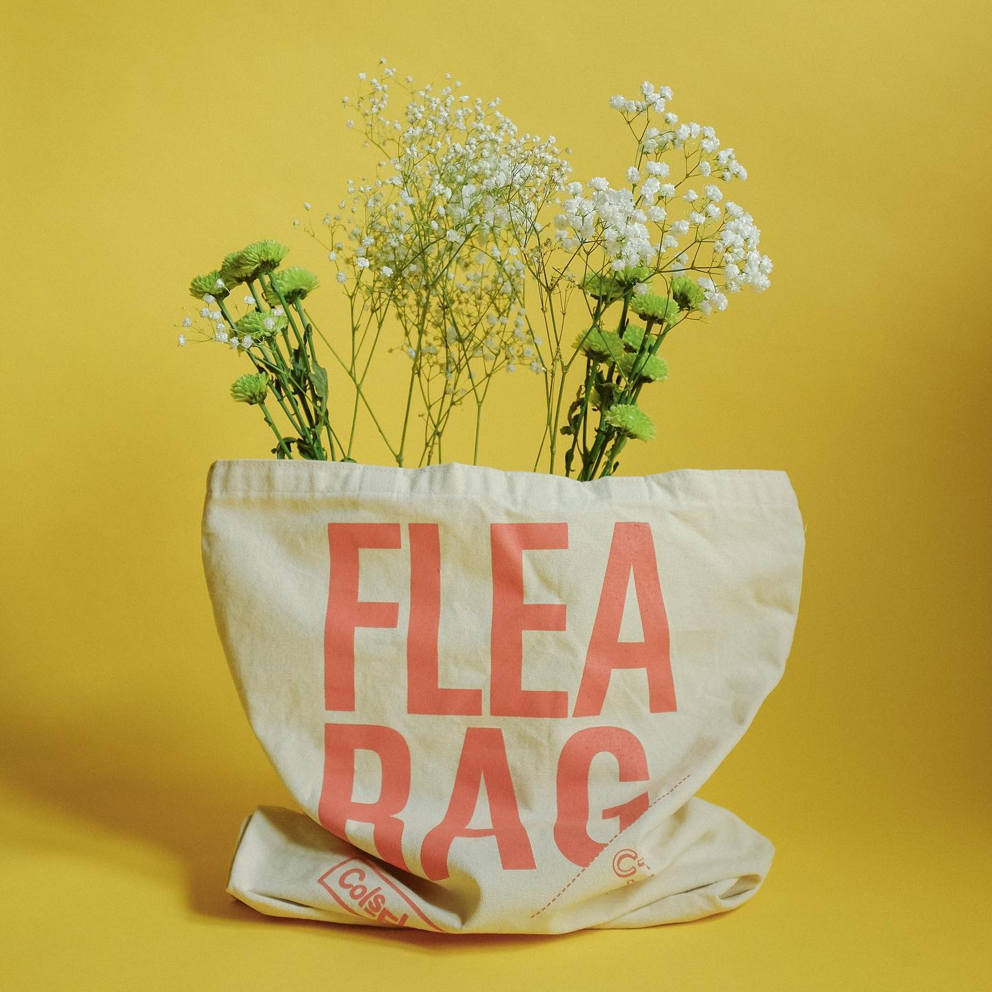 A canvas tote that reads "Flea Bag" standing upright with wildflowers extending out the top, against a bright yellow background