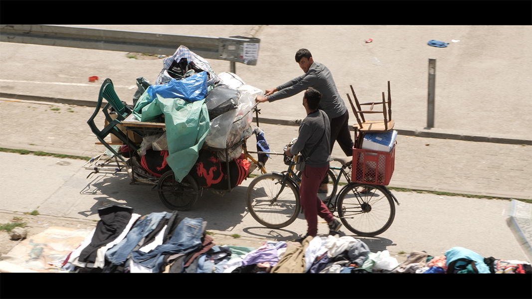 Two men on bikes push a cart laden with tarps, blankets, chairs, and other objects.