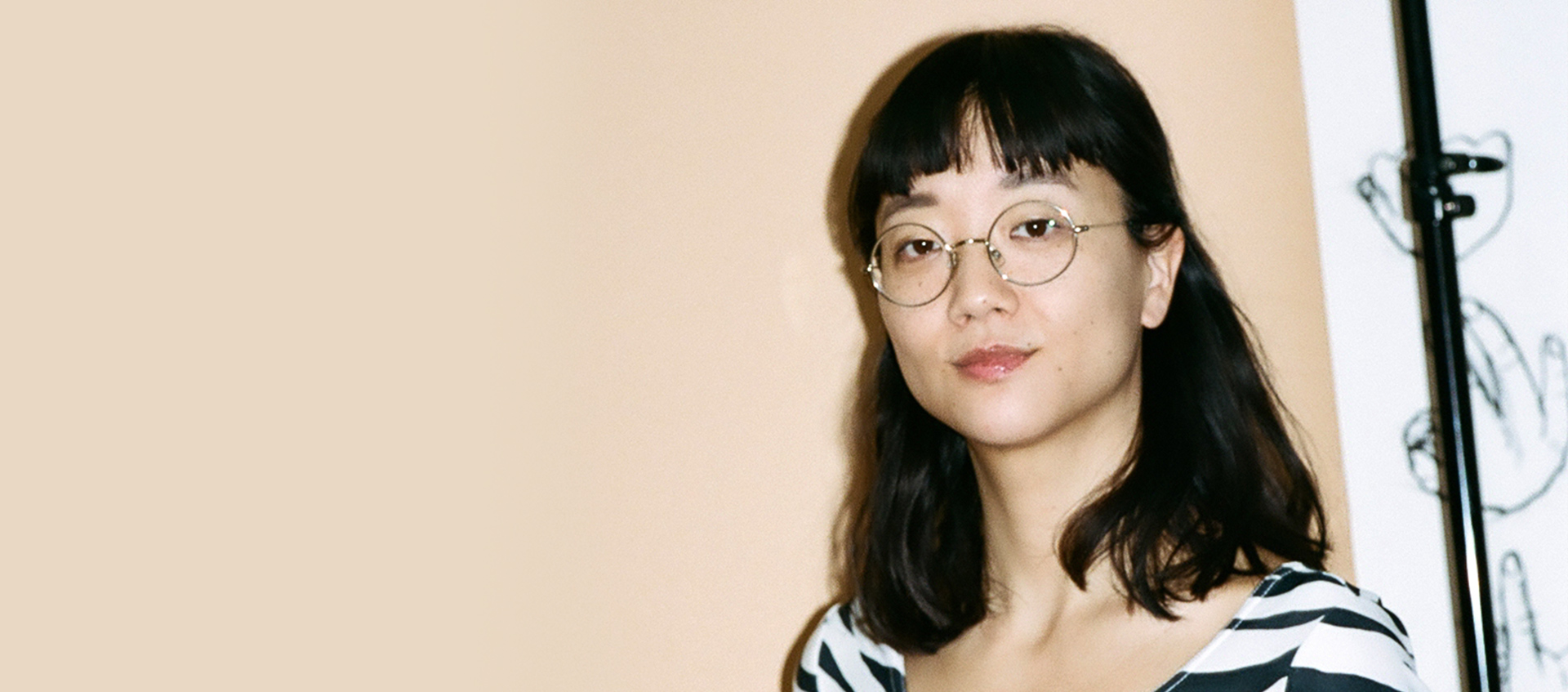 Photo of Christine Sun Kim in front of beige wall. Christine is wearing glasses and striped shirt