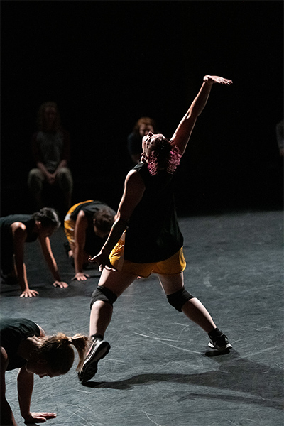 Dancer with back to the camera throwing their head back, with arm outstretched above their head. Three other dancers on the floor surrounding the dancer in center