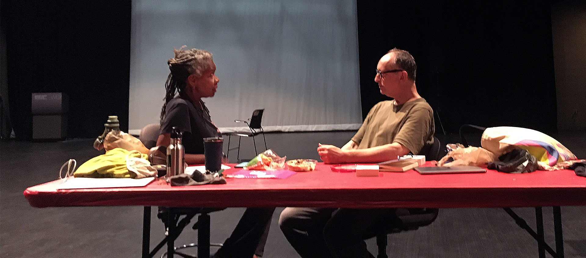 Bebe Miller (on left) and Paul Lazar (on right) seated and facing each other at a red table